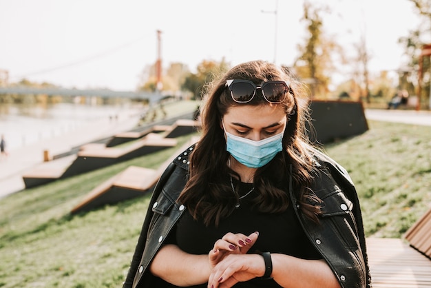 Großes Porträt einer schönen Frau in einer medizinischen Maske und Lederjacke in einem Park in der Natur 1