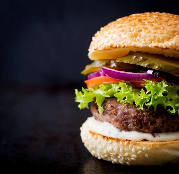 Großes Sandwich - Hamburgerburger mit Rindfleisch, Essiggurken, Tomate und Remoulade auf schwarzem Hintergrund.