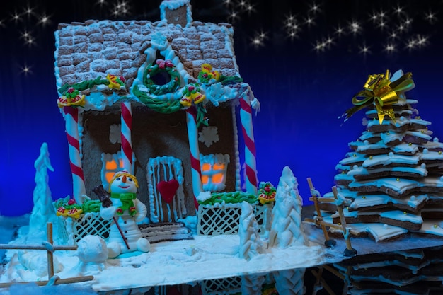 Großes schneebedecktes hausgemachtes Lebkuchenhaus mit Lichtern im Inneren, Lebkuchen-Weihnachtsbaum und einem Zuckermastix-Schneemann gegen Sternenhimmel. Mockup für saisonale Angebote und Urlaubspostkarte