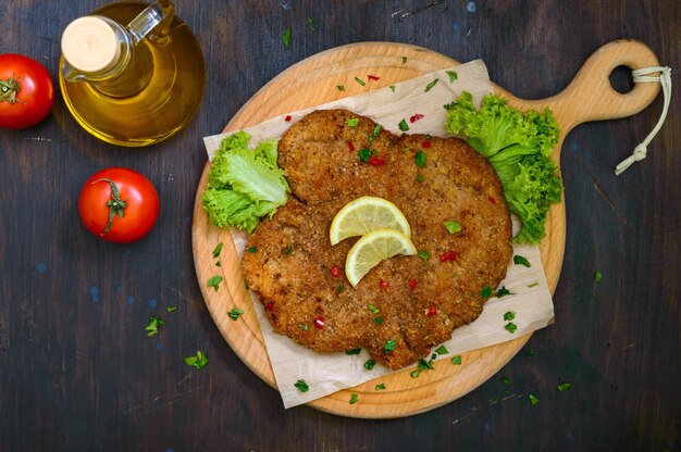 Großes Wiener Schnitzel auf einem Holzbrett mit Zitrone auf dunklem Hintergrund. Fleischgericht. Ansicht von oben, flach liegend.