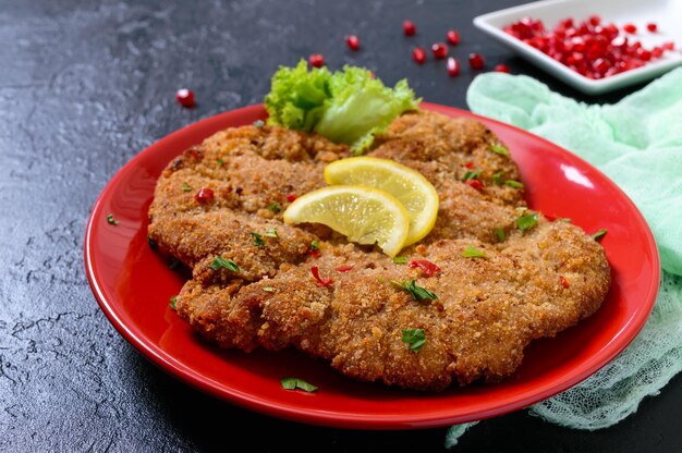 Großes Wiener Schnitzel auf einem roten Teller mit Zitrone auf schwarzem Hintergrund. Fleischgericht.