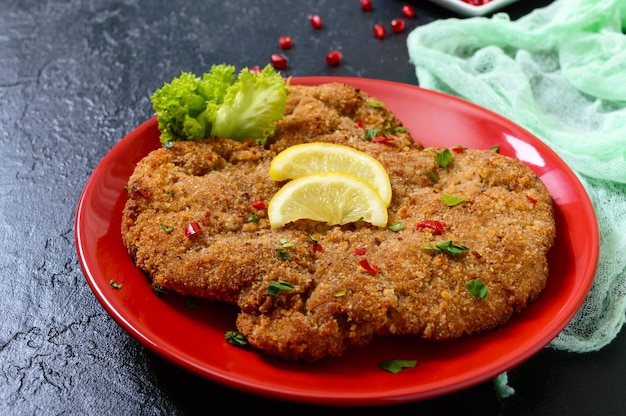 Großes Wiener Schnitzel auf einem roten Teller mit Zitrone auf schwarzem Hintergrund. Fleischgericht.