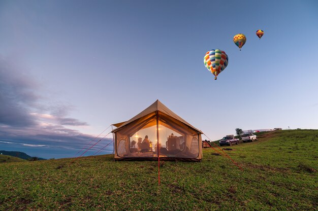 Großes Zelt mit Touristen, die auf grünem Hügel glühen, und Heißluftballons, die am Abendhimmel auf dem Land fliegen