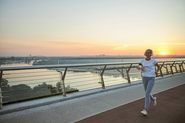 Großformatiges Porträt einer lächelnden Sportlerin mit einer Flasche Wasser, die über die Brücke läuft