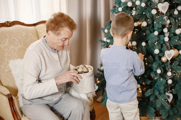 Großmutter mit ihrem Enkel am Weihnachtstag. Alte Frau und kleiner Junge schmücken den Weihnachtsbaum. Frau und Junge, die beige und blaue Kleidung tragen.