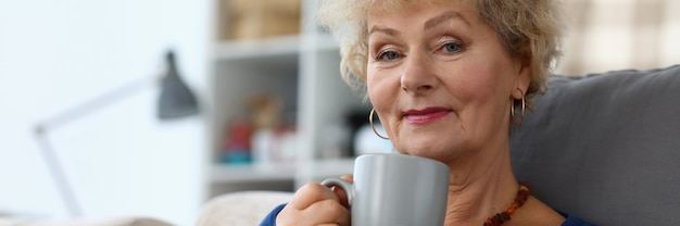 Großmutter trinkt Kaffee und liest Buch in der Wohnung. Schöne ältere Frau im Ruhestand sitzen zu Hause auf der Couch und ruhen sich aus.