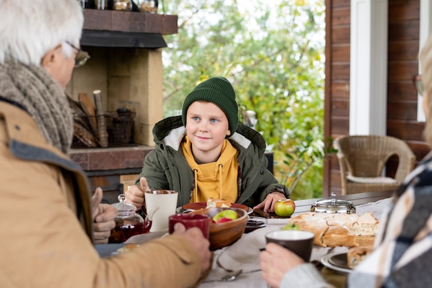 Großvater in warmer Freizeitkleidung und Wollstrickschal im Gespräch mit Enkel am bedienten Tisch beim Abendessen im Freien im Landhaus