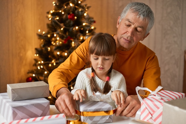 Großvater und Enkelin, die Geschenke verpacken, während sie am Tisch im Zimmer mit Weihnachtsbaum sitzen