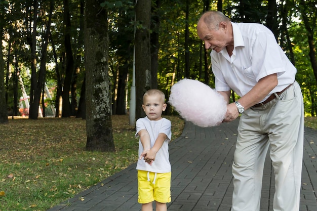 Großvater verwöhnt und füttert seinen kleinen Enkel liebevoll und sorgfältig mit rosa Zuckerwatte in einem Vergnügungspark Ein Rentner und ein kleiner Junge verbringen ihre Freizeit am Wochenende