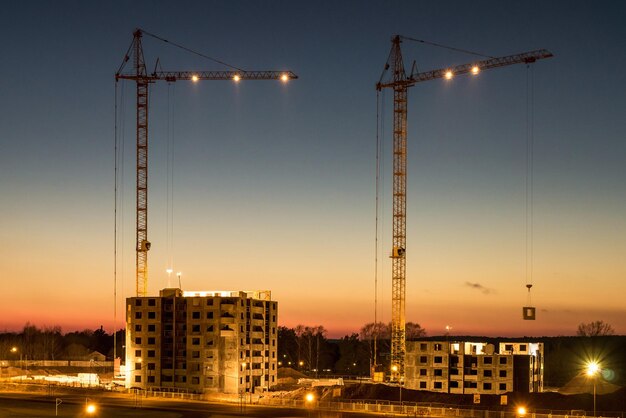 Grúas torre de silueta y edificios altos de varios pisos sin terminar en construcción al atardecer en el desierto en un sitio de construcción iluminado