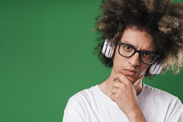 grübelnder junger Mann mit Afro-Frisur, der seinen Kopf stützt und Kopfhörer verwendet, die auf Grün isoliert sind