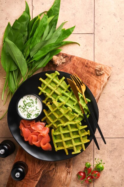 Grüne belgische Waffeln Spinat- oder Wildknoblauch- oder Pesto-Waffeln mit rotem Lachs und Sahnesauce auf altem beigefarbenem Fliesentischhintergrund Köstlicher Frühstücks-Snack-Brunch auf dunklem Hintergrund Draufsicht