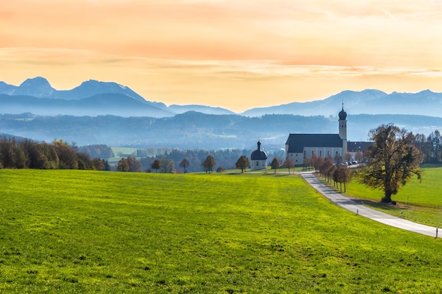 Grüne bunte Wiese, Straße, Kapelle, Kirche und Berge am nebligen Herbstmorgen. Alpenlandschaft, Bayerische Alpen, Deutschland.