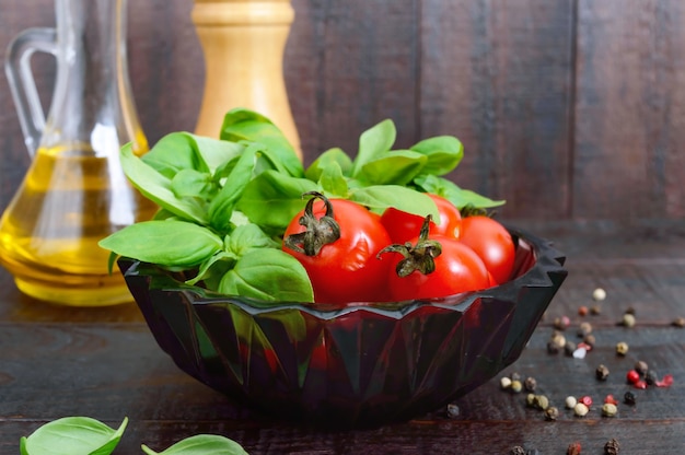 Grüne frische Blätter von Bio-Basilikum und kleinen reifen Tomaten, Öl und Pfeffer auf einem Holztisch für eine gesunde Ernährung.