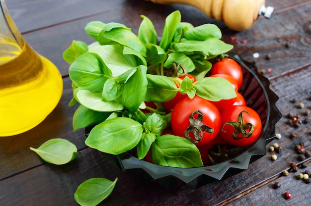 Grüne frische Blätter von Bio-Basilikum und kleinen reifen Tomaten, Öl und Pfeffer auf einem Holztisch für eine gesunde Ernährung.