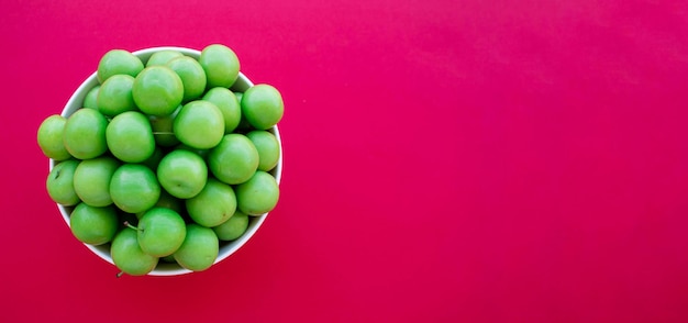 Grüne Pflaumen in der Schüssel lokalisiert auf rotem Hintergrund. Draufsichtfoto von sauren Pflaumenfrüchten.