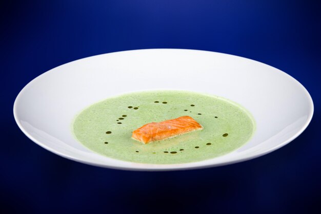 Grüne Spinatpüreesuppe mit Lachsscheiben. Flache Draufsicht auf eine weiße Platte auf einem blauen Hintergrund.