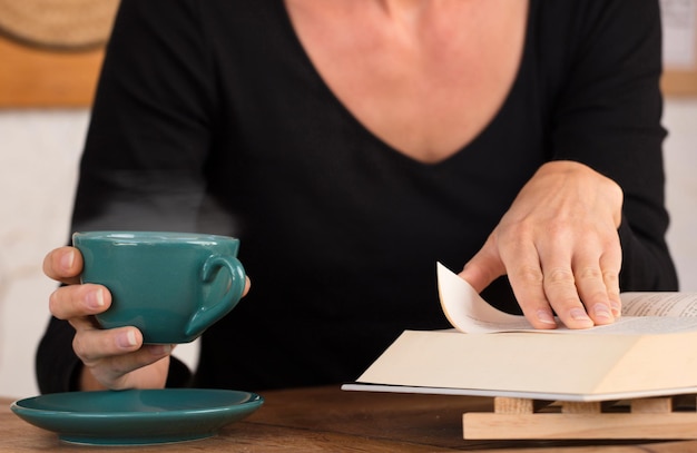 Grüne Tasse mit heißem Kaffee in den Händen der Frau mit Lesebuch zu Hause konzentrieren sich auf die Tasse