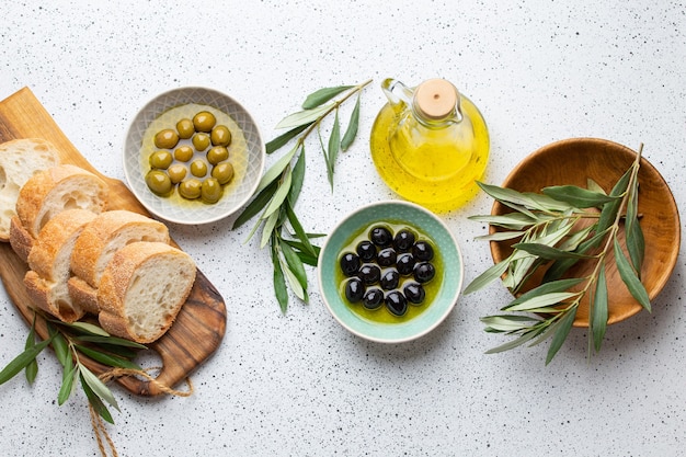 Grüne und schwarze Oliven mit Olivenöl in einer Glasflasche, Olivenbaumzweige und geschnittenes frisches Ciabatta-Brot auf Holzbrett. Weißer rustikaler Hintergrund, mediterranes Essenskonzept, Draufsicht