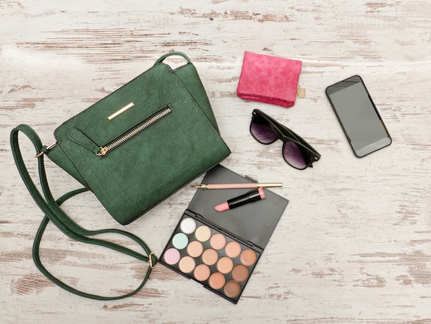 Grüne weibliche Tasche, Telefon, Lidschatten-Palette, Telefon, Sonnenbrille und Lippenstift auf einem hölzernen Hintergrund.