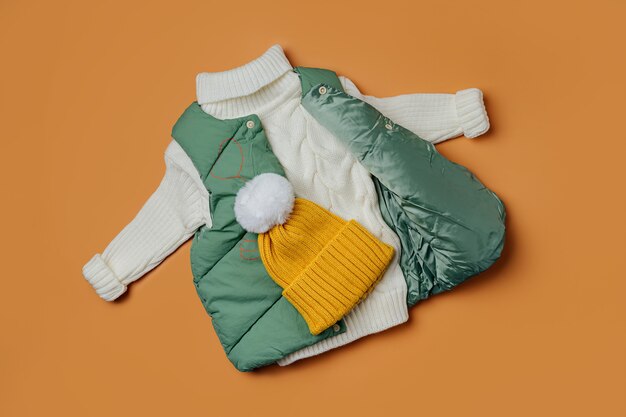 Grüne Westenjacken mit warmem Pullover auf orangem Hintergrund. Stylische Kinderoberbekleidung. Wintermode-Outfit