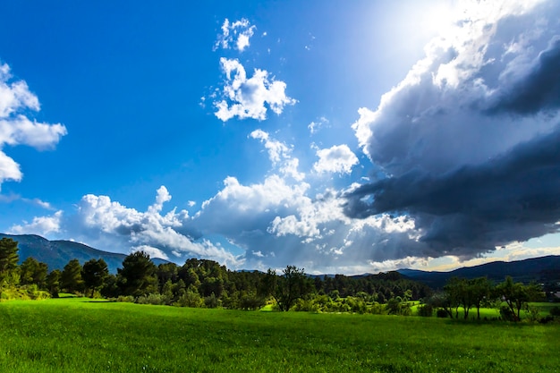 Grüne Wiese an einem Tag mit weißen und grauen Wolken und Sonnenstrahlen mit Bergen im Hintergrund.