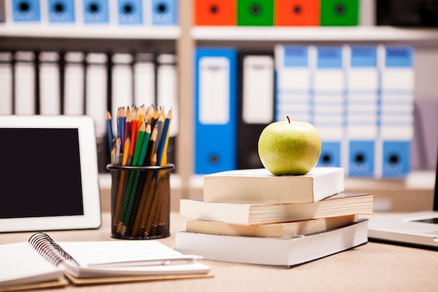 Grüner Apfel auf Bücherstapel neben einem Notizbuch und Bleistiften auf dem Tisch mit einem verschwommenen weißen Brett im Rücken. Schulkonzept