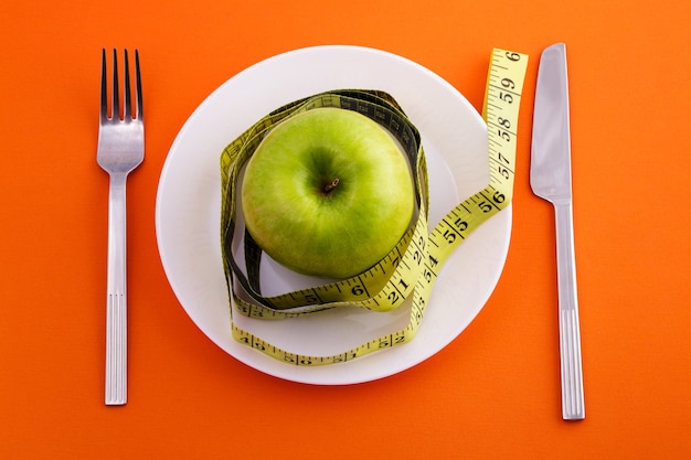 Grüner Apfel, der auf einem weißen Teller mit einem zurückgespulten Maßbandmesser und einer Gabel auf einer orangefarbenen Oberfläche liegt lying