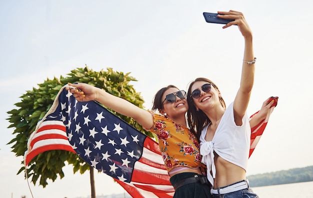 Grüner baum im hintergrund. zwei patriotische fröhliche frauen mit usa-flagge in den händen machen selfie im freien im park.