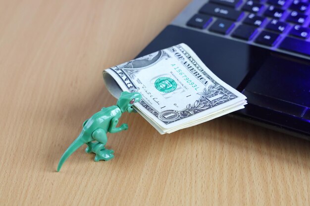 Grüner Dinosaurier trägt eine Packung Dollar zum Laptop im Mund, um das Konzept zu bezahlen, ist unerfahren