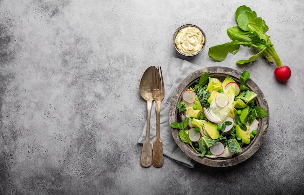 Grüner Gemüsesalat mit Avocado, Grünkohl, sprießenden Kräutern in rustikaler Schüssel serviert mit Sauce, grauer Betonhintergrund, Nahaufnahme, Draufsicht. Gesundes, sauberes Essen, Diät-/Detox-Konzept mit Platz für Text