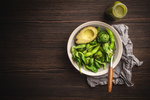 Grüner gesunder Salat mit Spinat, Rosenkohl, Avocado in Schüssel und grünem Detox-Smoothie in einer Flasche auf Holzhintergrund, Draufsicht. Rohkostdiät und sauberes veganes Essen, Platz für Text