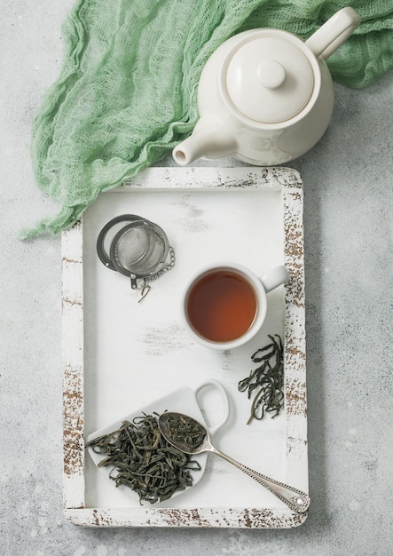 Grüner loser Tee mit Teekugelsieb und keramischer Teekanne mit Tasse in Holzkiste mit grünem Tuch auf hellem Hintergrund