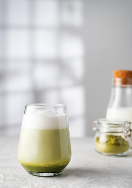 Grüner Matcha-Latte in einem Glas auf grauem Hintergrund mit morgendlichen Schatten aus dem Fenster eine Flasche Milch und Teepulver Das Konzept eines veganen Getränks, Superfoods und gesunder Lebensmittel Kopieren Sie Platz