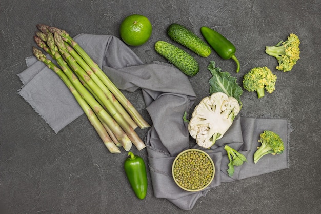 Grüner spargel, mungobohne und blumenkohl auf grauer serviette. paprika, gurken auf dem tisch. schwarzer hintergrund. flach legen