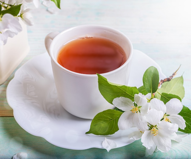 Grüner Tee in der weißen Tasse mit Zweig des blühenden Apfelbaums auf geformter Untertasse auf Holztisch. Nahansicht.