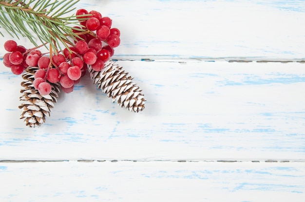 Grüner Zweig eines Weihnachtsbaumes mit einem Kegel und Gruppen von roten Beeren auf einer weißen hölzernen Hintergrundnahaufnahme