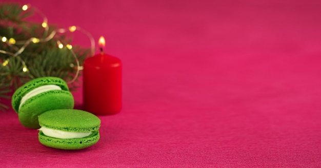 Grünes macaron mit Fondant auf rotem Papierhintergrund. In der Nähe befindet sich ein Weihnachtsbaumzweig mit einer Girlande und einer brennenden roten Kerze. Nahaufnahme, copyspace.