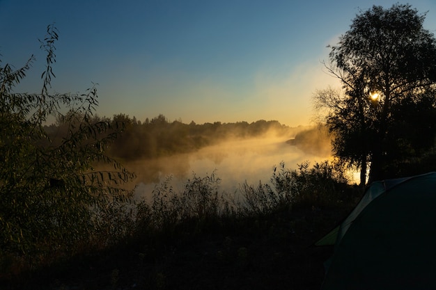 Grünes Touristenzelt am Fluss bei Sonnenaufgang, mit morgendlichem Herbstnebel auf dem Wasser. Touristische Landschaft im Freien.