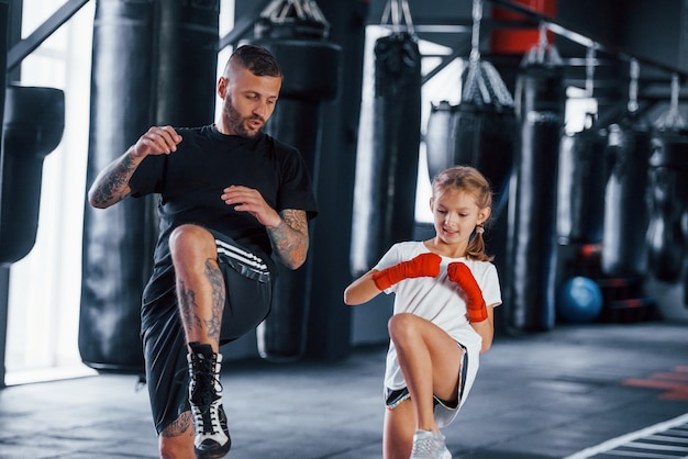 Grundschläge. Junger tätowierter Boxtrainer unterrichtet süßes kleines Mädchen in der Turnhalle.