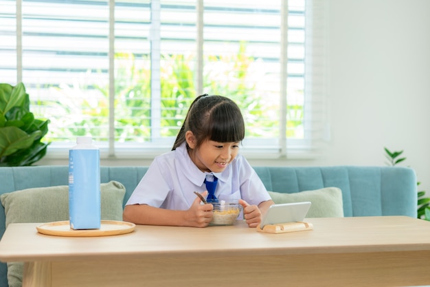 Grundschülerin in Uniform, die Frühstücksflocken mit Milch isst und Karikatur im Smartphone schaut