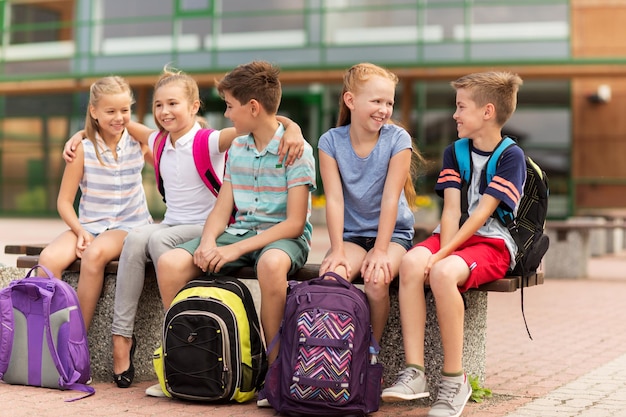 grundschulbildung, freundschaft, kindheit, kommunikation und menschenkonzept - gruppe glücklicher grundschüler mit rucksäcken, die auf einer bank sitzen und sich im freien unterhalten