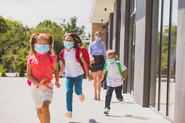 Grundschulkinder in medizinischen Masken, die nach dem Ende des langen Schultages im Freien laufen Junge Lehrerin sieht ihre Schüler ab, die während der Coronavirus-Pandemie in der Schule lernen