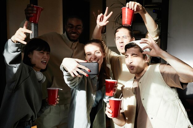 Grupo de adolescentes con bebidas posando en la cámara del teléfono móvil haciendo retrato selfie durante una fiesta