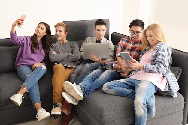 Grupo de adolescentes con dispositivos modernos que toman selfie mientras están sentados en un sofá en el interior