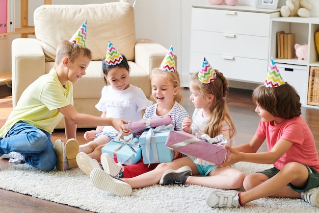 Grupo de adorables amigos en gorras de cumpleaños dando sus regalos a la feliz fiesta de niña rubia en casa