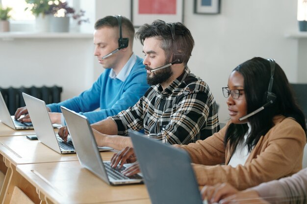 Grupo de agentes de call center multiétnicos jóvenes concentrados sentados en mesas y usando computadoras portátiles mientras venden productos por teléfono