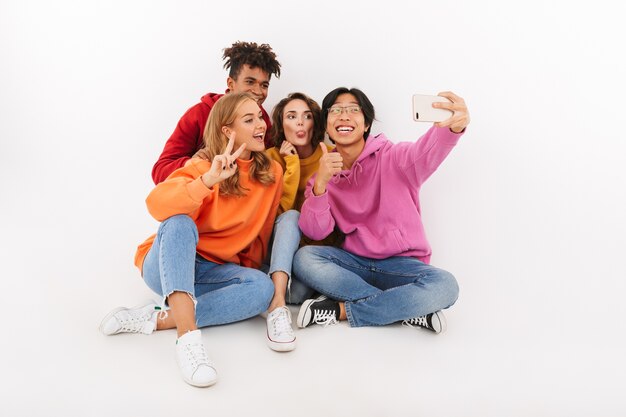 Grupo de alegres amigos multirraciales sentados aislados, tomando un selfie con teléfono móvil