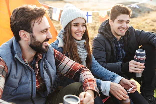 Grupo de alegres jóvenes amigos acampando al aire libre, sentados con tazas y termo