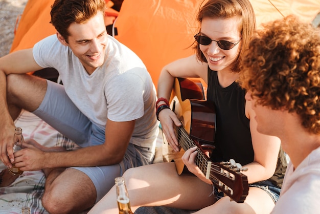 Grupo de alegres jóvenes amigos divirtiéndose juntos en la playa, bebiendo cerveza, tocando la guitarra mientras acampa